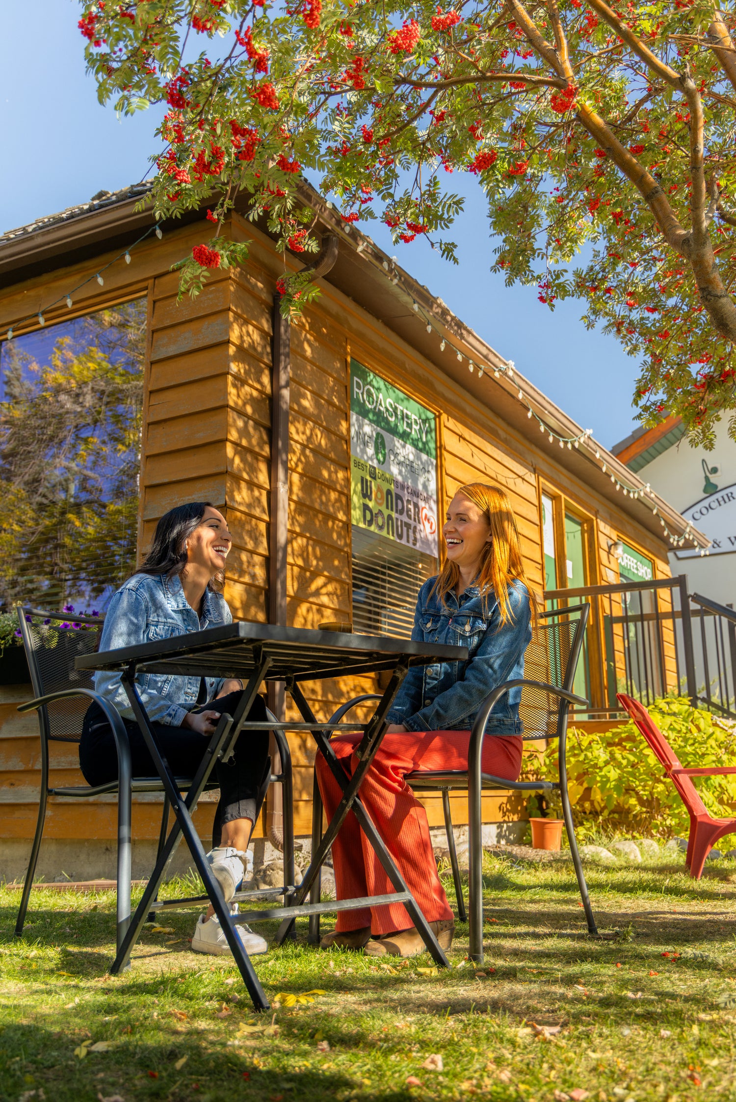 Two women sitting at a table outdoors in front of a wooden building Photo by JRNY Media